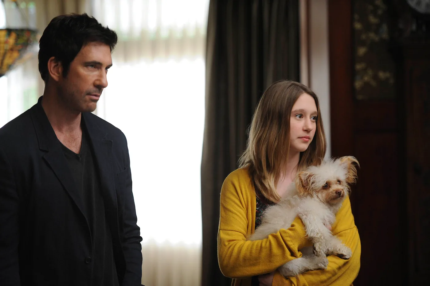 Dylan McDermott as Ben Harmon stands next to Taissa Farmiga as Violet, who holds a small white dog, in a Season 1 publicity still.