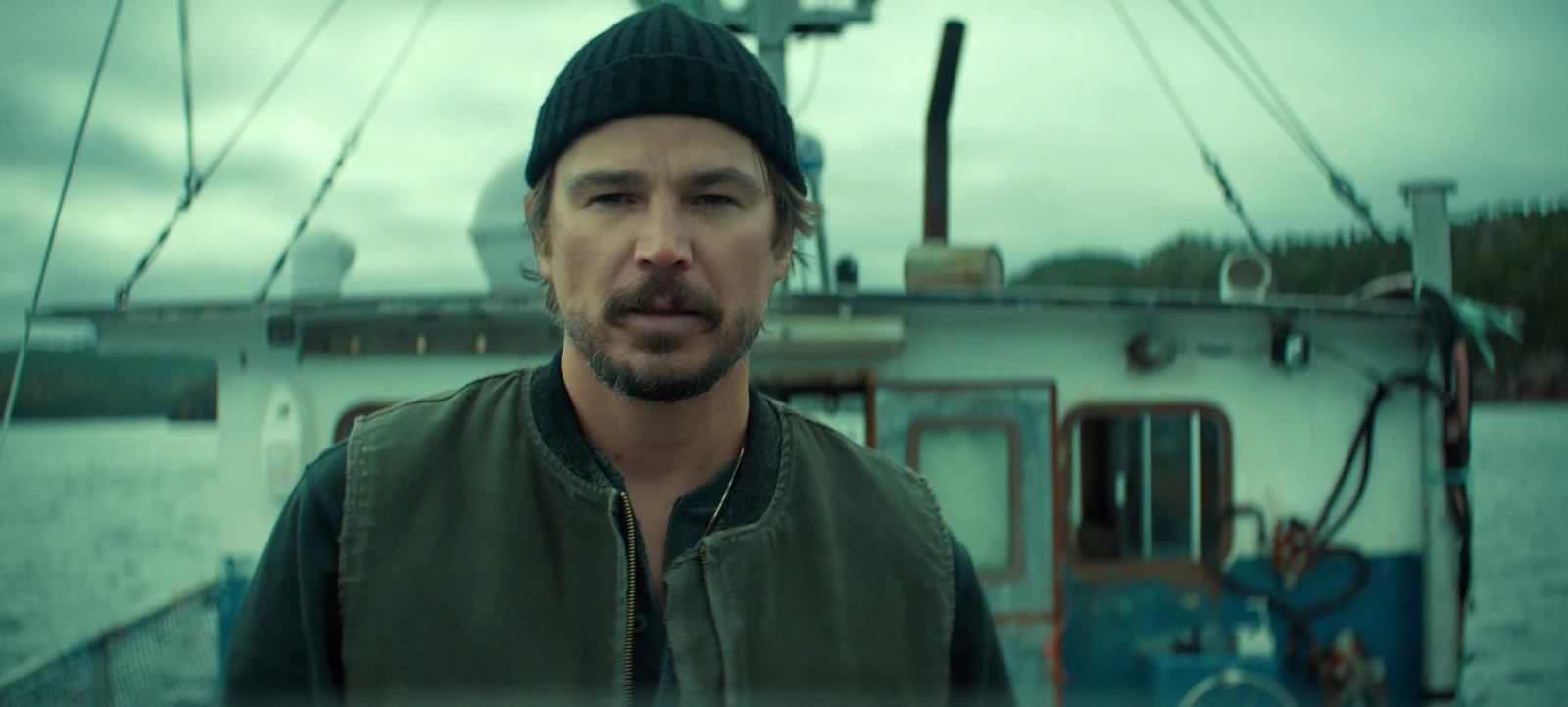 Josh Hartnett as Calvin Penney standing on a fishing boat in Netflix's Below, wearing a black beanie and green vest against an overcast Newfoundland harbor.