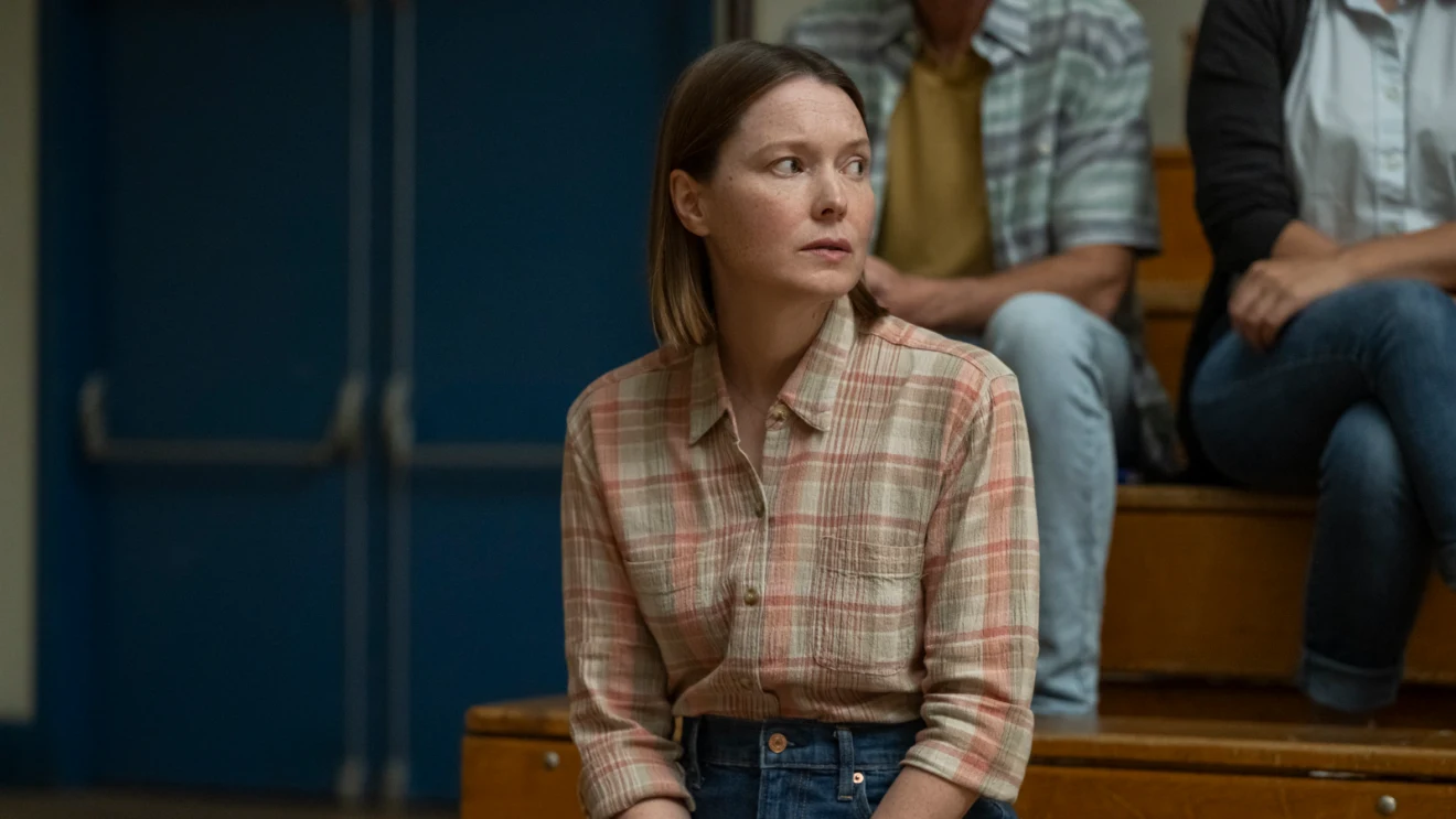 Samantha Sloyan as Margaret White sitting on the wooden bleachers of a school gymnasium.
