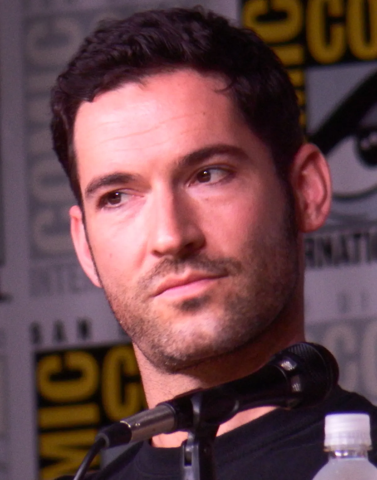 Tom Ellis in a dark polo shirt seated behind a microphone at San Diego Comic-Con in 2016.
