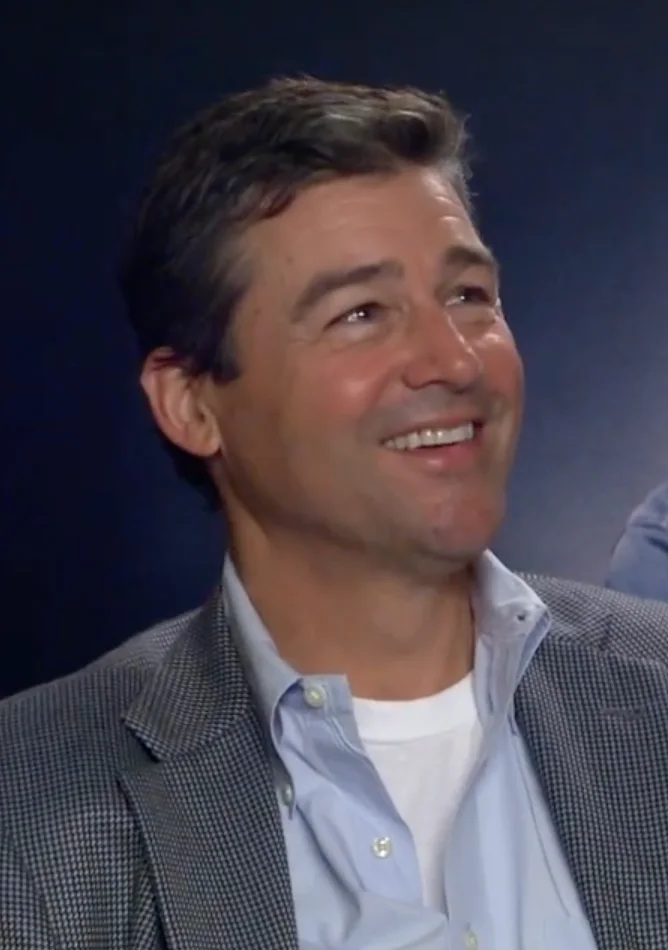 Kyle Chandler smiles while looking to one side, wearing a grey patterned blazer over a light-blue shirt.