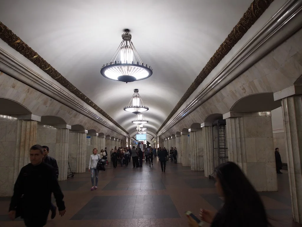 Commuters walk through a Moscow Metro station hall lined with stone columns and chandeliers.