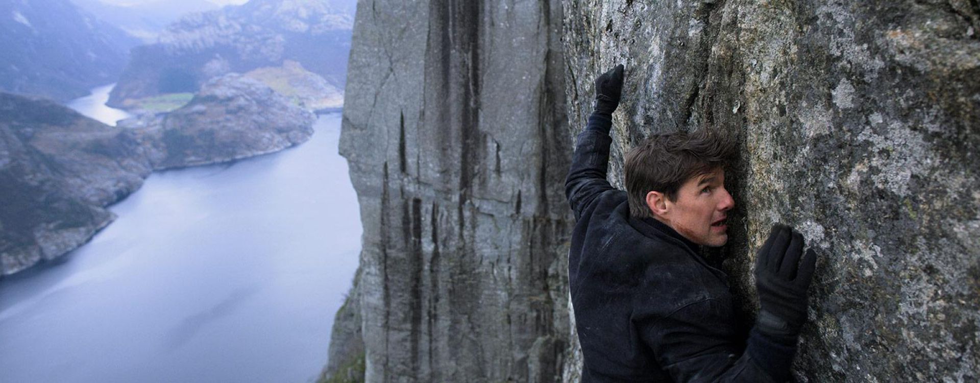 Tom Cruise as Ethan Hunt clings to a cliff face in a Mission: Impossible – Fallout promotional still.
